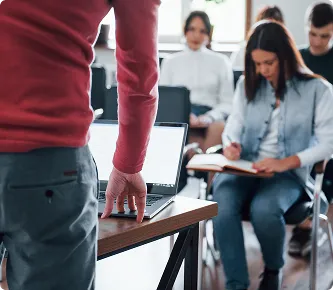 Formador de pie ante un portátil con asistentes sentados tomando notas